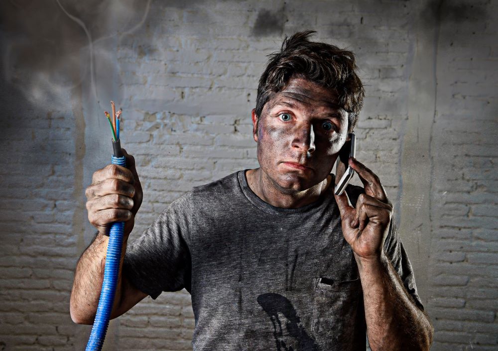 young man with a dirty burnt face is holding a smoking electrical cable in one hand and a phone on the other