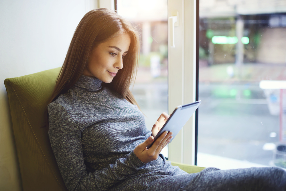 Portrait of young beautiful female relaxing in cafe interior sitting near window while reading electronic book and enjoying silents using modern touchpad and free wireless connection in wifi zone