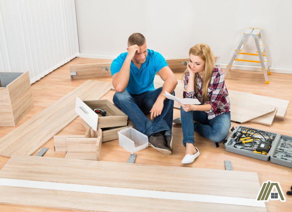 Man and woman sitting on the floor looking at the instructions manual to assemble a big furniture