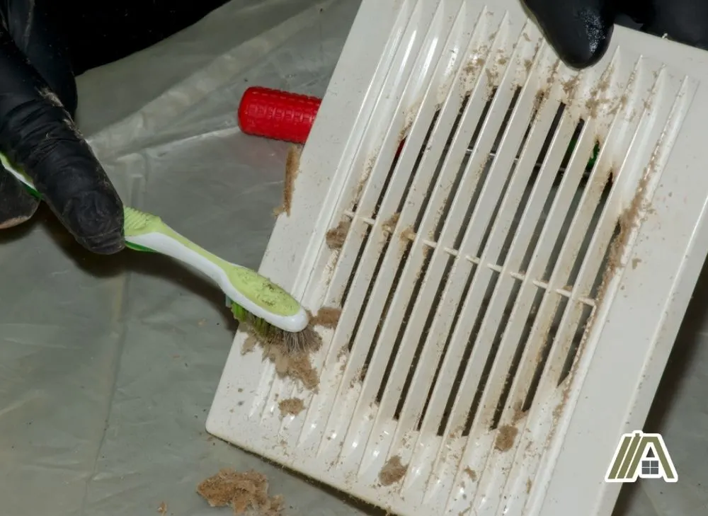 Man wearing gloves while cleaning the bathroom fan cover using a toothbrush